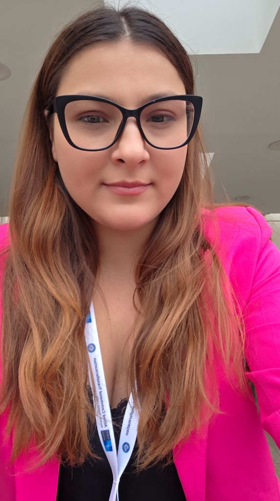 Femeie cu părul lung, cămașă neagră și sacou roz, purtând ochelari, zâmbind în fața camerei.